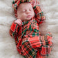 A sleeping newborn baby wearing a red, green, and yellow plaid footed onesie with a matching plaid bow headband, lying on a white faux fur blanket.