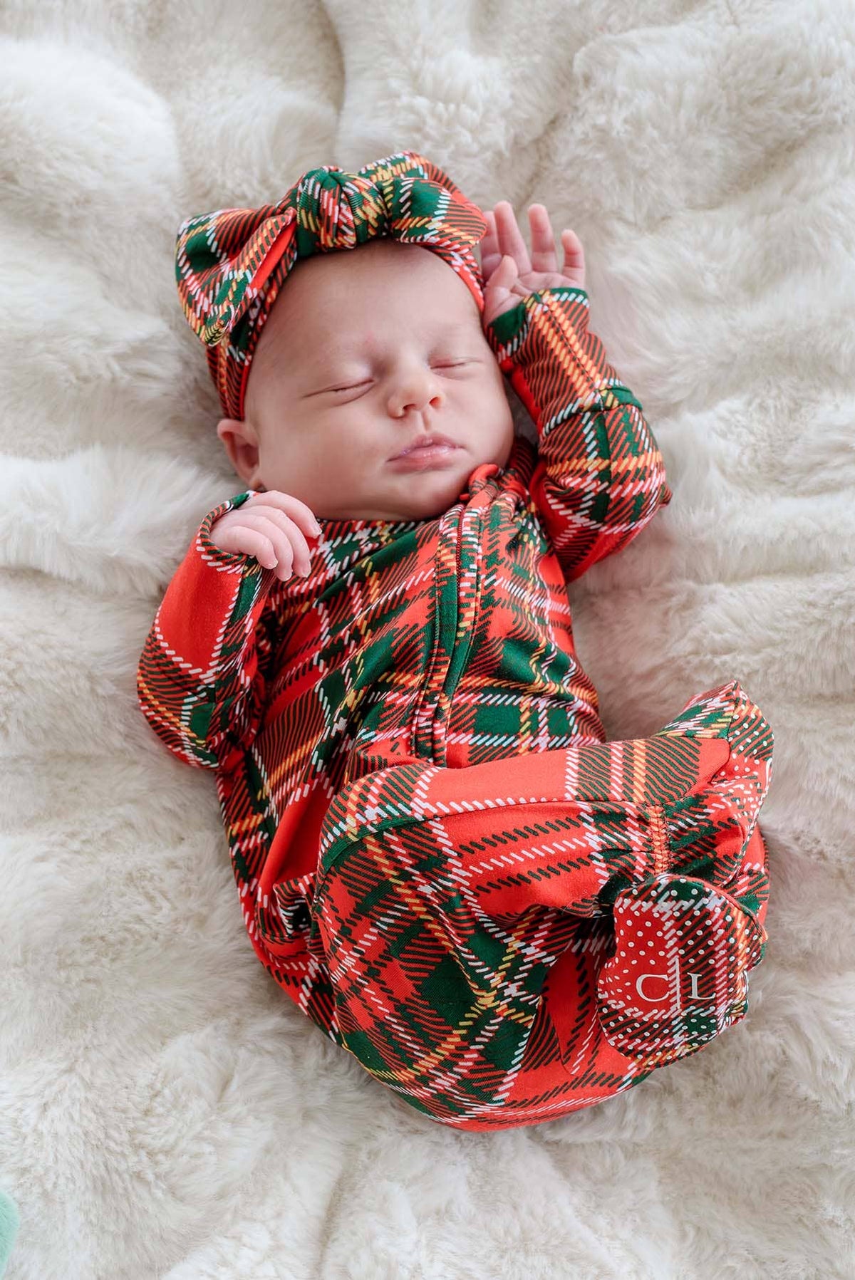 A sleeping newborn baby wearing a red, green, and yellow plaid footed onesie with a matching plaid bow headband, lying on a white faux fur blanket.