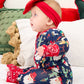 A baby wearing a festive Christmas-themed navy blue one-piece jumpsuit with Santa Claus and holiday prints, paired with a matching red bow headband, sitting on a white bed with holiday pillows.