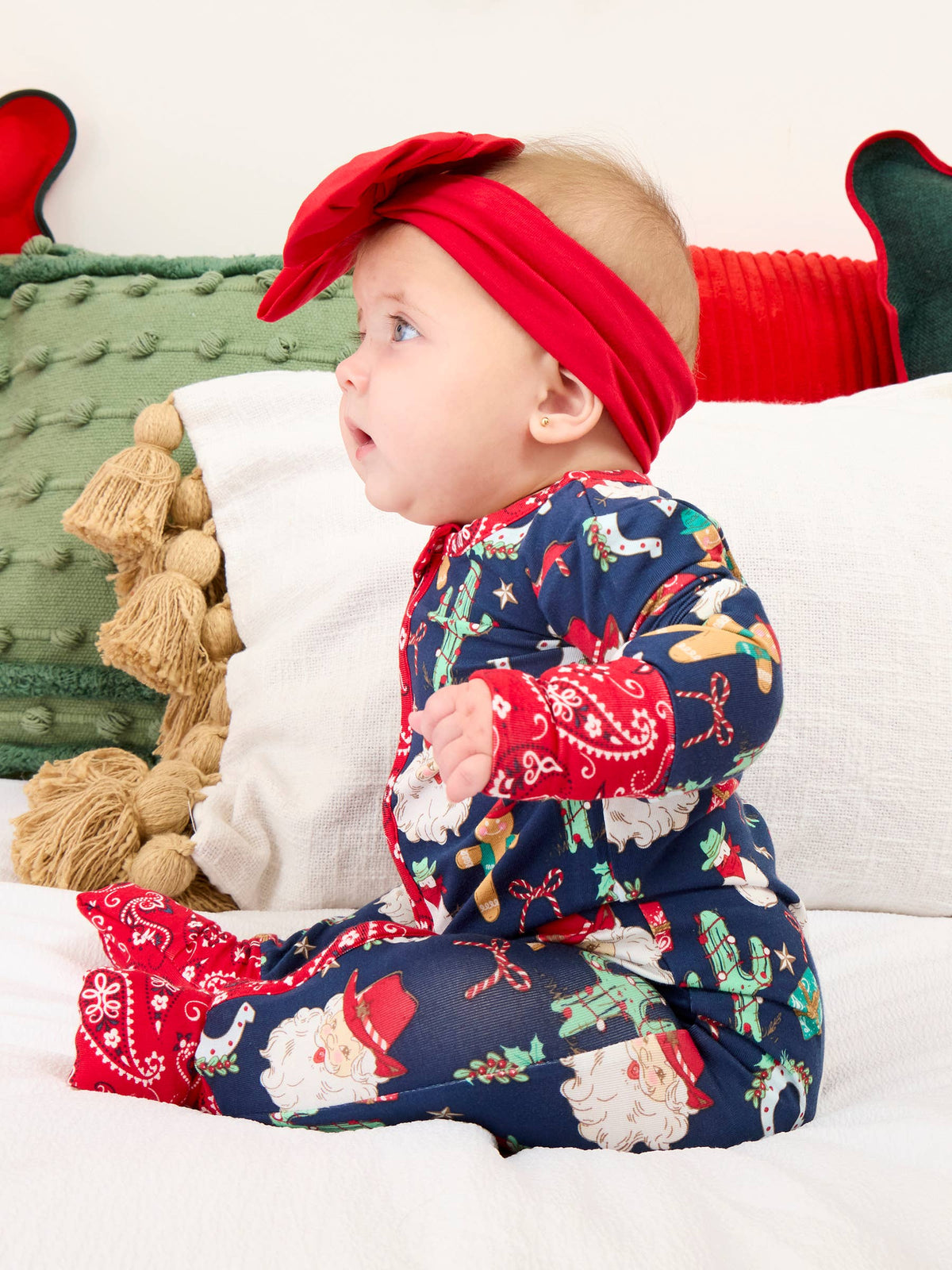 A baby wearing a festive Christmas-themed navy blue one-piece jumpsuit with Santa Claus and holiday prints, paired with a matching red bow headband, sitting on a white bed with holiday pillows.