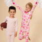 Two children wearing long-sleeve pajamas pose against a solid beige background. The child on the left wears a white pajama set with small orange polka dots and holds a brown football.