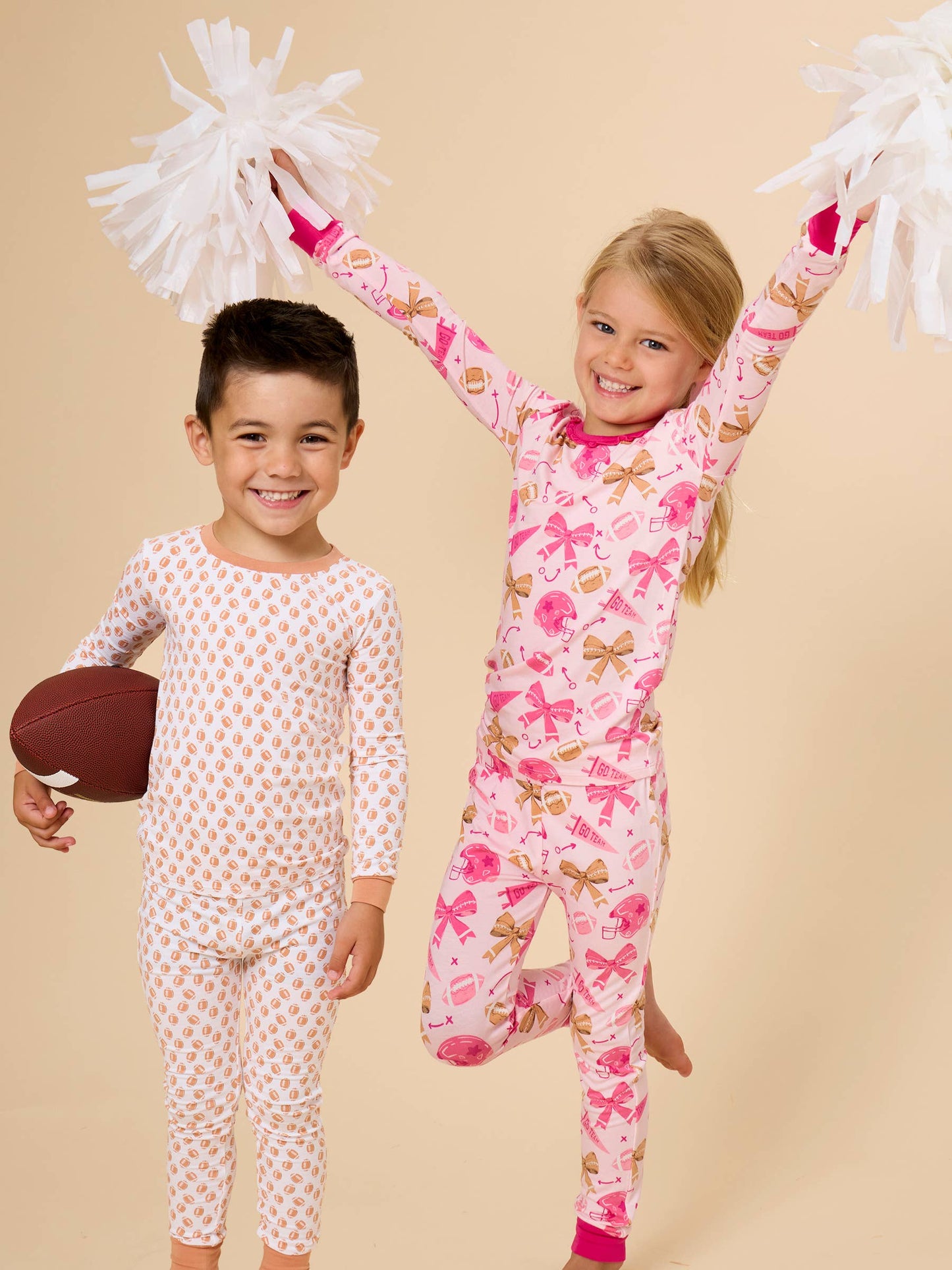 Two children wearing long-sleeve pajamas pose against a solid beige background. The child on the left wears a white pajama set with small orange polka dots and holds a brown football.
