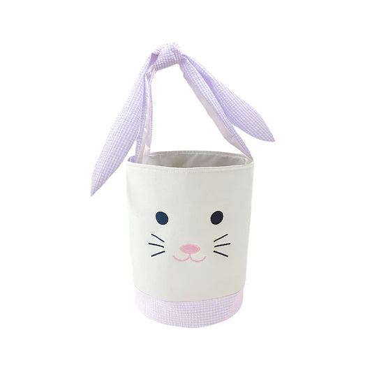 A white cotton tote basket with lavender trim, featuring a embroidered bunny face and bunny ear straps.