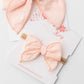 Two pale peach-pink bow hair accessories displayed on branded white cards. One larger bow is draped on the top card, and a smaller bow on a wooden stick is attached to the lower card.