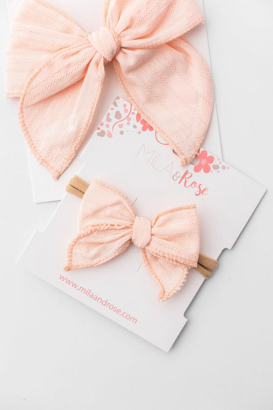 Two pale peach-pink bow hair accessories displayed on branded white cards. One larger bow is draped on the top card, and a smaller bow on a wooden stick is attached to the lower card.