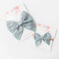 Two sage hair bows on white cards with floral designs.