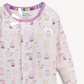 A close-up of a baby long-sleeve pajama top with a white base and a colorful pattern of illustrated milk bottles, jars, and desserts. The pajama has a purple collar and a visible interior neckband with rainbow stripes.
