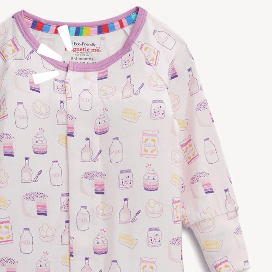 A close-up of a baby long-sleeve pajama top with a white base and a colorful pattern of illustrated milk bottles, jars, and desserts. The pajama has a purple collar and a visible interior neckband with rainbow stripes.
