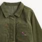 Close-up of an olive green corduroy jacket with a pointed collar, two front chest pockets, orange contrast stitching, and a small embroidered squirrel motif on the left chest pocket.