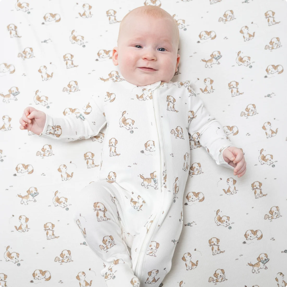A baby lying on their back wearing a white footed zip-up sleeper with a brown and beige dog print, set against a matching dog-patterned blanket.