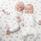 Two smiling babies lying side by side on a white fitted sheet with a brown dog print, wearing matching white footless zip-up sleeper outfits. One baby has a pink headband.