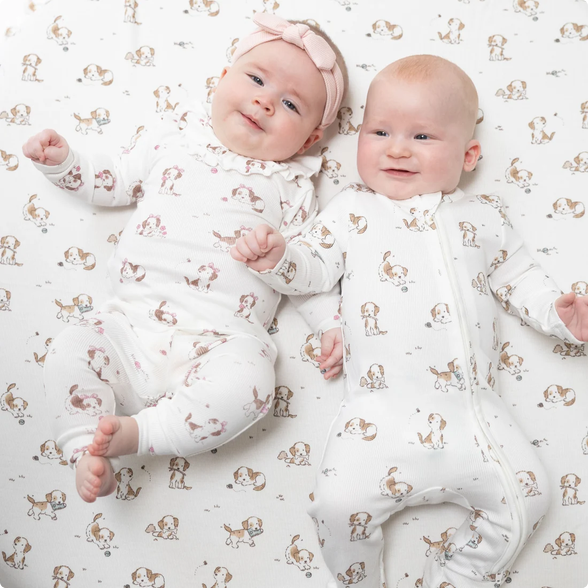 Two smiling babies lying side by side on a white fitted sheet with a brown dog print, wearing matching white footless zip-up sleeper outfits. One baby has a pink headband.