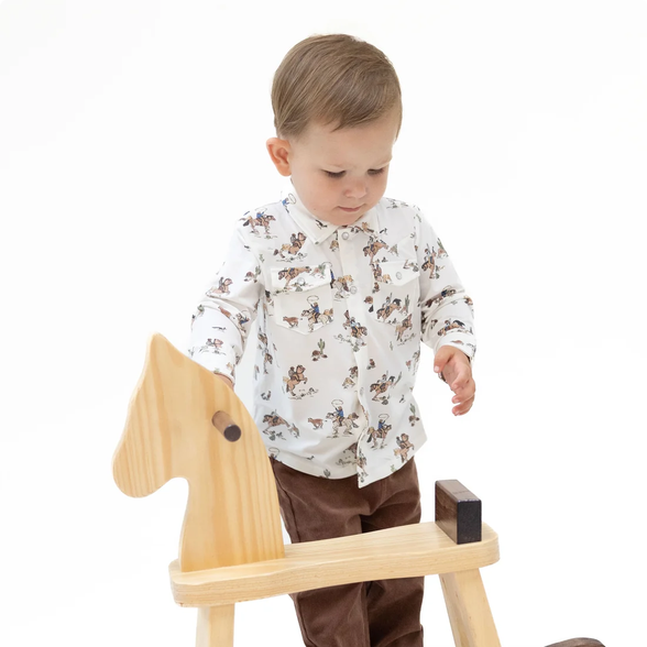 A toddler boy wearing a white long-sleeve button-up shirt with a brown horse pattern and brown pants, standing behind a light wood rocking horse toy on a white background.