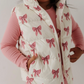 A young child wearing a cream quilted puffer vest with a pink bow print, layered over a pink ribbed long-sleeve top and pink leggings, shown against a plain light background.