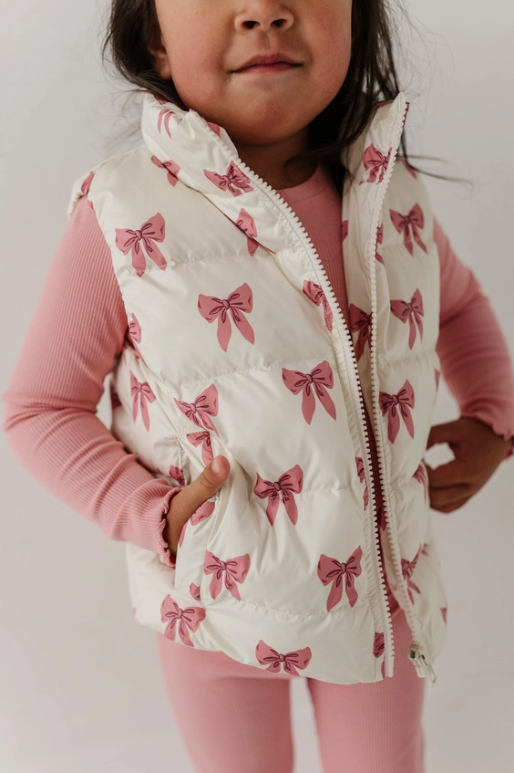 A young child wearing a cream quilted puffer vest with a pink bow print, layered over a pink ribbed long-sleeve top and pink leggings, shown against a plain light background.