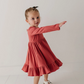 A young girl with blonde hair in a small bow is wearing a long-sleeve terracotta pink tiered dress and white Mary Jane shoes, posing with one arm extended against a plain white studio background.