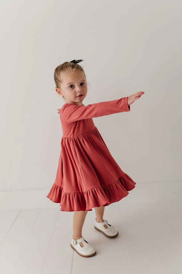 A young girl with blonde hair in a small bow is wearing a long-sleeve terracotta pink tiered dress and white Mary Jane shoes, posing with one arm extended against a plain white studio background.