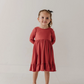A young girl stands against a plain white studio background wearing a long-sleeve terracotta-colored dress with a ruffled hem and white shoes.