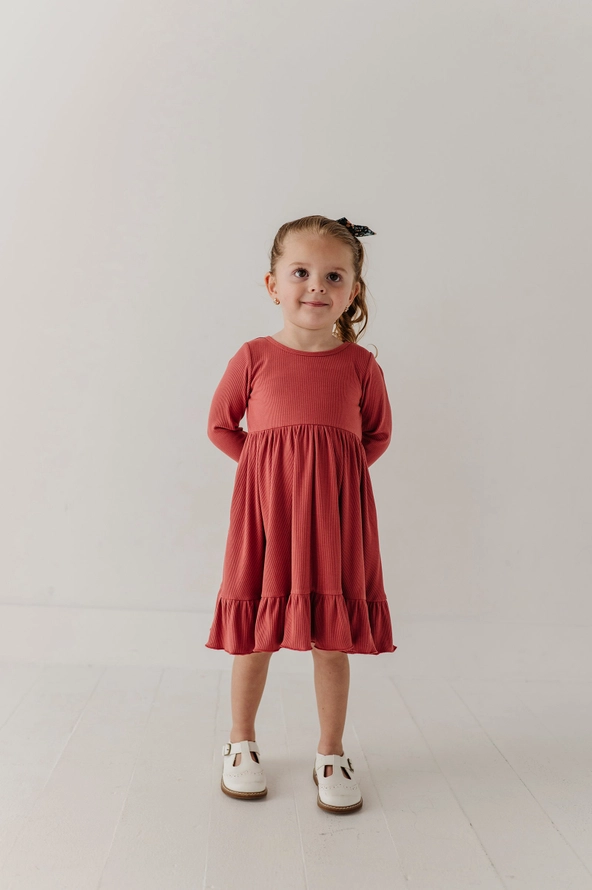 A young girl stands against a plain white studio background wearing a long-sleeve terracotta-colored dress with a ruffled hem and white shoes.