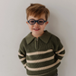 A young boy with short brown hair and round black glasses, wearing an olive green and cream striped knit polo sweater and gray jeans, standing against a plain light gray wall.