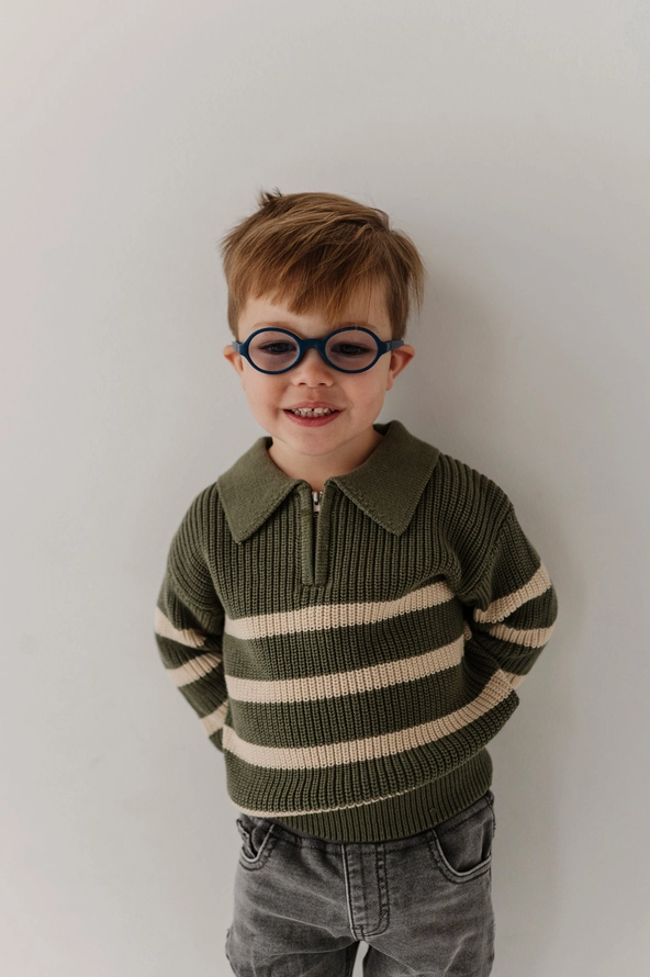 A young boy with short brown hair and round black glasses, wearing an olive green and cream striped knit polo sweater and gray jeans, standing against a plain light gray wall.