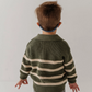 A toddler viewed from behind wearing an olive green and cream striped knit sweater, gray jeans, and a blue patterned headband against a plain white studio background.