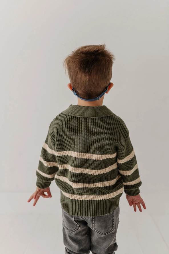 A toddler viewed from behind wearing an olive green and cream striped knit sweater, gray jeans, and a blue patterned headband against a plain white studio background.