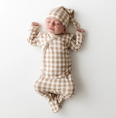 A sleeping newborn baby lying on their back, wearing a beige and white gingham check long-sleeve footed sleeper and matching beanie hat.