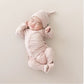 A sleeping newborn baby wearing a light pink long-sleeve footed sleeper and matching pointed hat, lying on a neutral beige background.