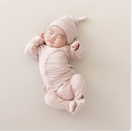 A sleeping newborn baby wearing a light pink long-sleeve footed sleeper and matching pointed hat, lying on a neutral beige background.