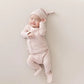 A sleeping newborn baby wearing a light pink long-sleeve footed bodysuit and matching pointed sleep hat, lying on a neutral beige background.