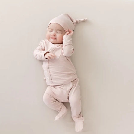 A sleeping newborn baby wearing a light pink long-sleeve footed bodysuit and matching pointed sleep hat, lying on a neutral beige background.