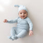 A smiling baby lying on their back wearing a light blue long-sleeve footed sleeper and matching pointed sleep hat against a plain light beige background.