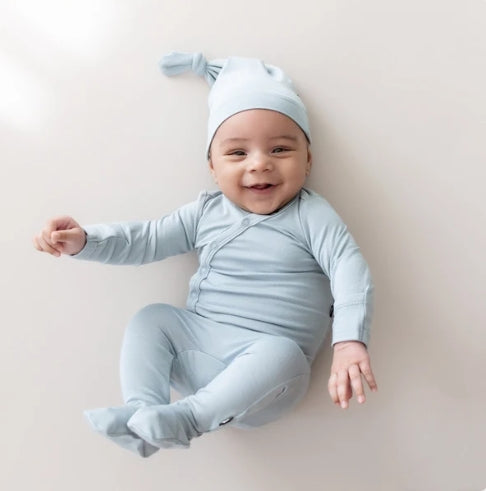 A smiling baby lying on their back wearing a light blue long-sleeve footed sleeper and matching pointed sleep hat against a plain light beige background.