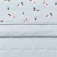 A close-up of a quilted baby blanket or comforter in a pale blue-gray color, featuring a top border printed with illustrated ducks and small reeds on a light blue background.