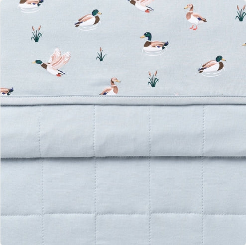 A close-up of a quilted baby blanket or comforter in a pale blue-gray color, featuring a top border printed with illustrated ducks and small reeds on a light blue background.