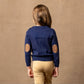 A child viewed from behind wearing a navy blue knit sweater with circular tan elbow patches and light beige pants, standing against a textured warm brown backdrop.