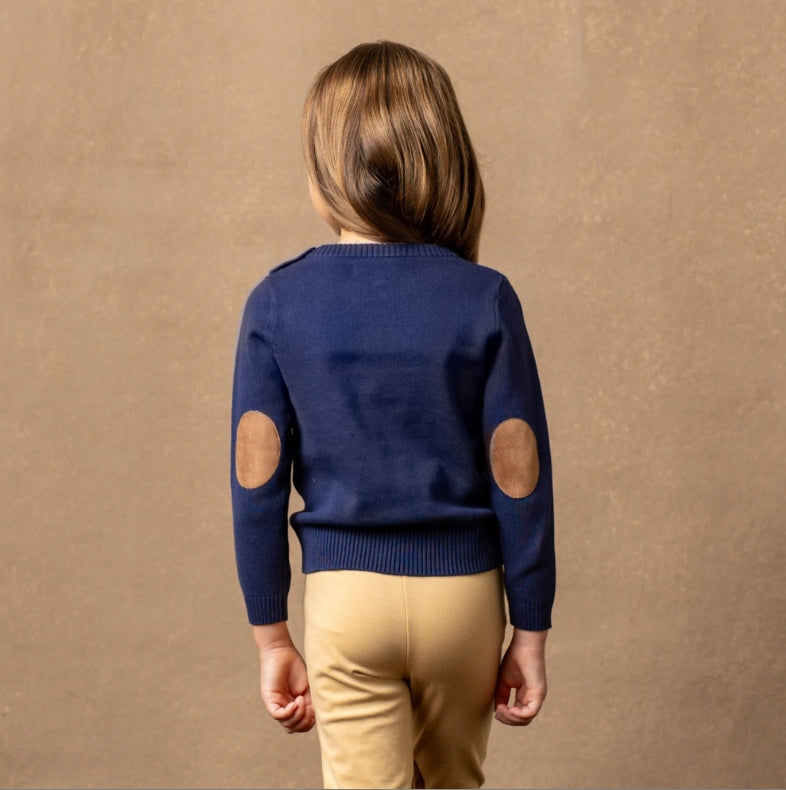 A child viewed from behind wearing a navy blue knit sweater with circular tan elbow patches and light beige pants, standing against a textured warm brown backdrop.
