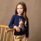 A young girl with long light brown hair stands indoors wearing a navy blue knit sweater with a large embroidered horse head motif on the front, paired with tan pants. She is posed next to a light wood chair against a warm brown textured backdrop.