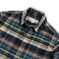 A close-up of a plaid button-down shirt collar and upper chest area. The fabric pattern features navy blue, green, yellow, and white intersecting plaid lines.