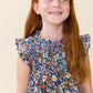 A young girl with long red hair wearing a blue floral smocked dress, smiling and looking to the side against a plain cream background.