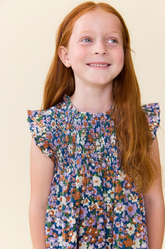 A young girl with long red hair wearing a blue floral smocked dress, smiling and looking to the side against a plain cream background.