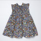 A sleeveless baby dress with a smocked bodice and ruffled shoulder straps, featuring an all-over dense floral print in blue, purple, orange, and white, laid flat on a light gray background.