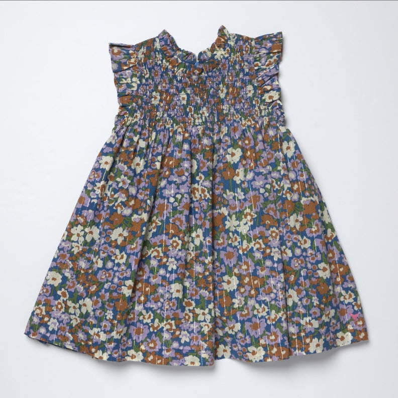 A sleeveless baby dress with a smocked bodice and ruffled shoulder straps, featuring an all-over dense floral print in blue, purple, orange, and white, laid flat on a light gray background.
