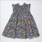 A sleeveless smocked dress with a dense multicolor floral print, featuring ruffled shoulder details and a gathered skirt, laid flat against a light gray background.