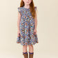 A young girl with long red hair stands against a plain beige studio background wearing a short-sleeve floral dress with ruffled shoulders and brown boots.