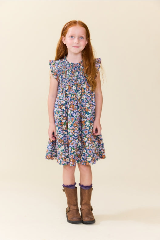A young girl with long red hair stands against a plain beige studio background wearing a short-sleeve floral dress with ruffled shoulders and brown boots.