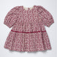 A short-sleeve baby dress with a dusty rose and cream floral block print pattern, featuring a smocked waist and a thin matching fabric waistband.