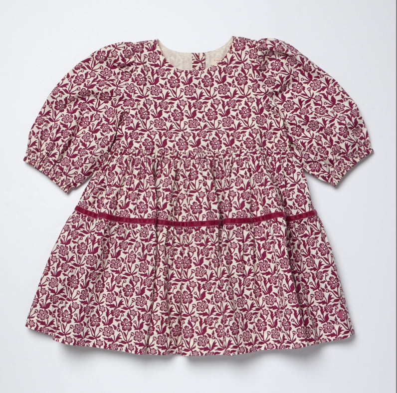 A short-sleeve baby dress with a dusty rose and cream floral block print pattern, featuring a smocked waist and a thin matching fabric waistband.