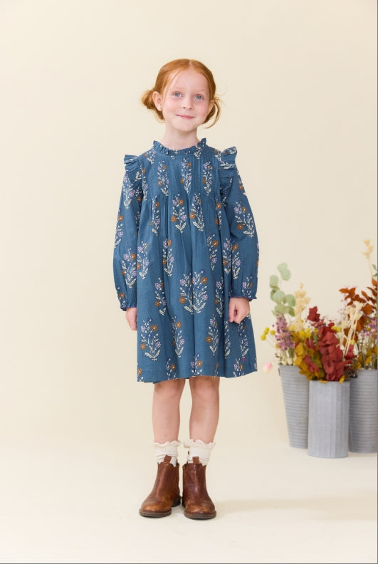 A young girl with red hair stands wearing a blue long-sleeve dress with a white floral pattern, white ruffled socks, and brown boots. She is posed in front of a plain beige background with dried flowers in grey pots on the right.